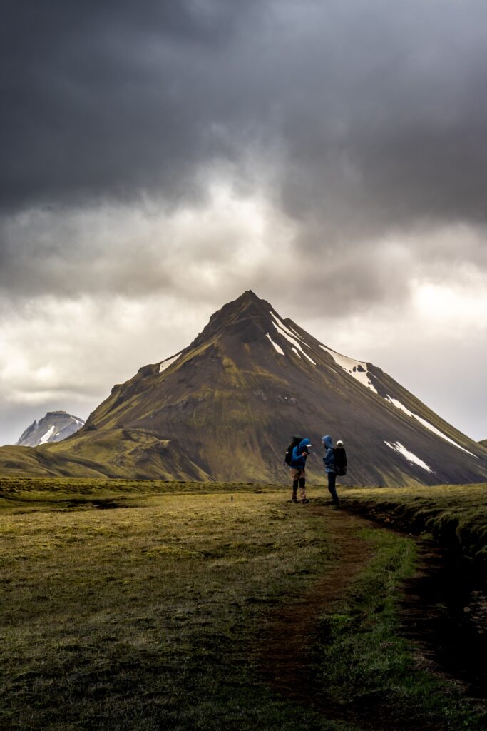 Two hikers moving along a mountain trail under dramatic storm clouds, illustrating danger, survival, and uncertain outcomes.